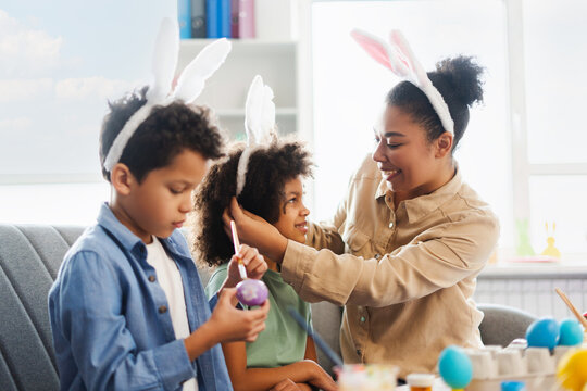 Smiling mother and cute kids wearing bunny ears painting and decorating easter eggs at home. Happy African American family celebration Easter together