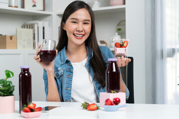 Smiling young beautiful Asian reviewing tasty mixed strawberry and cherry juice detox drinks,...