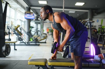Fototapeta na wymiar Young African American man sitting and lifting a dumbbell close to the rack at gym