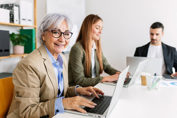Fototapeta na wymiar Smiling gray haired 60s senior businesswoman looking at camera sitting at workplace with young adult colleagues. Teamwork and business people concept.