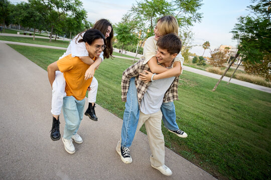 Cheerful multiracial couple doing a race in piggyback. Group of people laughing and having fun together outdoor garden area. Young students enjoying leisure and free time with friends in the park