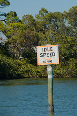 Idle speed sign orange and white on wood post in blue water on a sunny day. Blue sky and green trees in the background. Vertical shot with no people.