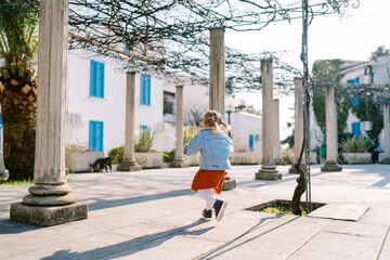 Little girl walks between pergola columns in a garden near a three-story building