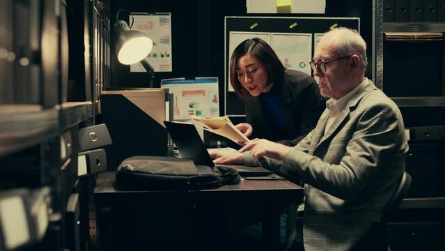 Law agents studying old classified information from police archives folders, looking for clues and crime trends. Partners accessing additional archived resources in incident room. Camera B.