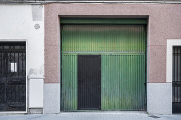 Large green metal doors of a garage and another small black access door in a ground floor premises with a security lock on the cement and brick facade