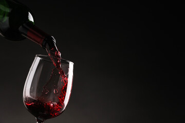 Pouring tasty red wine from bottle into glass on dark background, closeup. Space for text