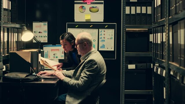 Law agents studying old classified information from police archives folders, looking for clues and crime trends. Partners accessing additional archived resources in incident room. Camera A.