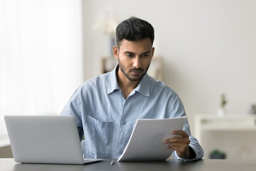 Indian man prepare agreement, review data working on laptop looks serious and focused busy in workflow sit at desk with wireless computer, read terms, check conditions, learn contract details document