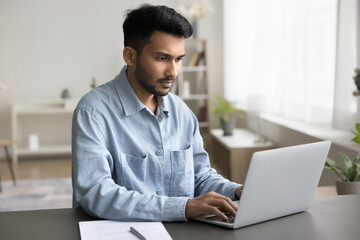 Serious Indian man sit at desk with laptop, work on online project, check email, search information...