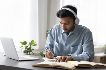 Indian man studying on-line on laptop, make notes in copybook, prepare essay, doing university assignment, learning at home using modern tech and audio course app. E-learning, new skills, education