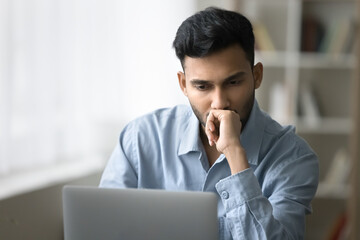 Serious concentrated Indian businessman staring at laptop screen think over business issue, read...