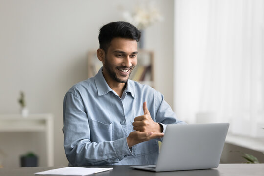 Young smiling Indian man with hearing disability sit at desk with laptop, make video call, take part in virtual meeting or online class showing hands gestures, lead communication using sign language
