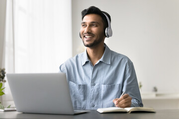 Happy Indian man in headphones make notes, listen audio course, improve foreign language skills,...