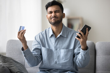 Young Indian man hold card and smartphone try to pay via e-bank feels angry, having problems with...