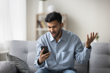 Indian guy staring at smartphone screen with angry face expression, get awful news in notice from bank, bad message, scam, fraud, screams due to short battery life, lagging phone, mobile apps crash - obrazy, fototapety, plakaty
