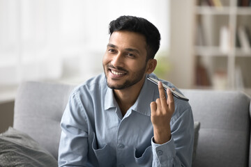 Smiling young Indian man hold smartphone near ear enjoy pleasant audio message, listening voicemail, exchanges sms by mobile application, get positive news, lead friendly conversation on speakerphone