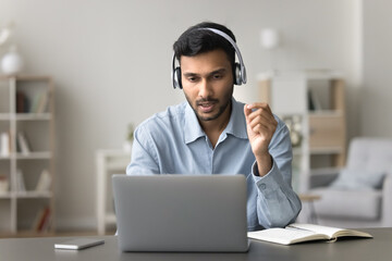 Young 30s Indian man in headphones sit at desk lead business negotiation remotely, talk to client...