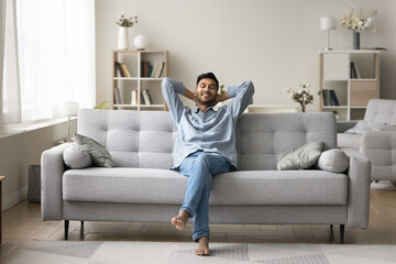Carefree wellbeing barefoot Indian guy enjoy fresh conditioned air leaned on comfortable sofa in...