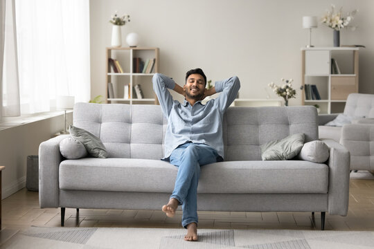 Carefree wellbeing barefoot Indian guy enjoy fresh conditioned air leaned on comfortable sofa in fashionable living room, relaxing alone with hands behind head. Rest, leisure, daydreams, smart home