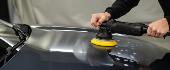 A mechanic polishes the surface of the hood of a gray car. 