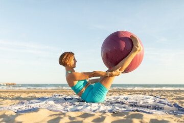 A woman practices the double-leg stretch movement on the beach while holding a Pilates ball