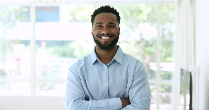 Confident handsome young African man in office shirt looking at camera with arms crossed, posing for head shot portrait indoors with large window in background, getting happy, cheerful, smiling