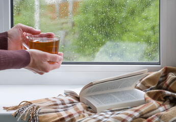 A woman's hands with a cup of tea. An open book and a soft blanket. The window glass is covered with raindrops. Springtime