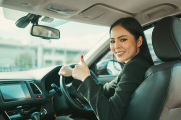 Young beautiful asian business women in suit getting new car showing thumbs up. She very happy and excited. Smiling female driving vehicle on the road on a bright day.