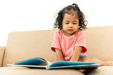 Focused little cute girl reading a book story on sofa in living room at home. Kids activity learning. Emotional, Cognitive development concept.