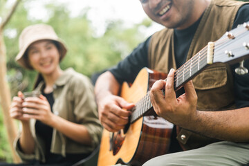 Young asian couple man and woman picnic sitting in campsite outdoor garden. Two people camping in forest playing guitar and drinking coffee. Travel relax camping on vacation holiday weekend theme