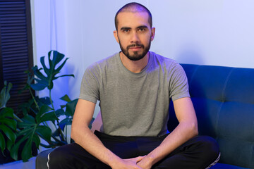 Fototapeta premium A short-haired man sitting cross-legged on a blue couch looking at the camera. He is Wearing a grey t-shirt and black pants. A indoor plant in the background and blue light illuminates the place.
