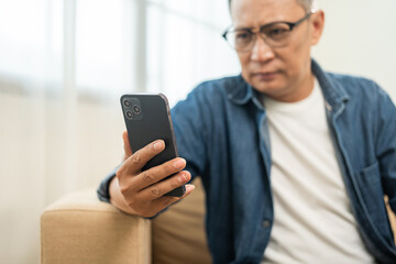 Obraz na płótnie Canvas Depressed Asian mature old man using smartphone serious thinking on sofa in living room. Portrait stress of senior asian man holding cell phone. Mature People and lifestyle