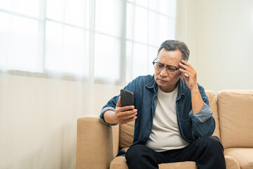 Depressed Asian mature old man using smartphone serious thinking on sofa in living room. Portrait stress of senior asian man holding cell phone. Mature People and lifestyle