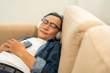 Mature lifestyle relaxing on sofa in the morning. Happy Asian senior old man listening music with smartphone put on headphone by streaming music online. Choosing song by cell phone
