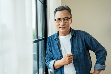 Handsome asian mature old man standing near windows with sunlight in the morning. Happy Portrait of cheerful smiling senior asian man pose looking at camera. Mature People and lifestyle - obrazy, fototapety, plakaty