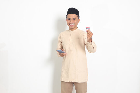 Portrait of attractive Asian muslim man in koko shirt with skullcap holding a mobile phone and presenting credit card. Isolated image on white background