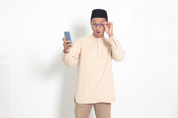 Portrait of surprised Asian muslim man in koko shirt with skullcap holding mobile phone, showing wow shocked face expression. Advertising and social media concept. Isolated image on white background