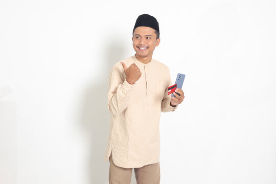 Portrait of attractive Asian muslim man in koko shirt with skullcap holding a mobile phone and credit card while pointing with finger to the side. Isolated image on white background