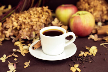 White coffee cup with espresso, cinnamon, red apples and dry hydrangea on dark brown background....