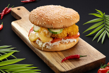 Delicious burger with fried chicken, cucumbers, cheese, tomatoes, onions and vegetables on a wooden board with pepper.Dark background. Food photos. Advertising for the menu.