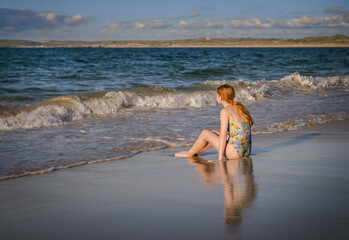 Teenage girl with red hair wearing swimming suit sitting on sandy beach at water's age looking at sea waves; distant shore and sky in background