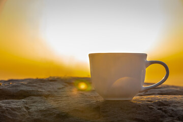 A white cup of hot espresso coffee mugs on the rocks and nature view of the sun background in the...