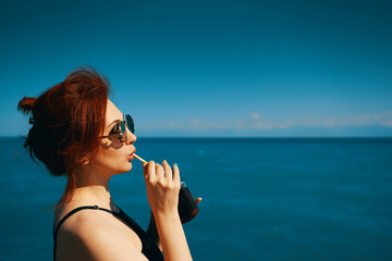 Woman on summer vacation. Red-haired girl in sunglasses drinks lemonade from bottle through cocktail tube. Blue sea, sunlight and clear sky on background. Copy space for travel agency advertising.