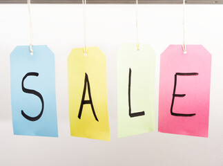 Closeup of four different color hanging tags or labels, each attached by a piece of string, and with a letter written in marker pen, spelling out the word SALE.