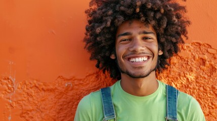 Young man with curly hair smiling broadly wearing a green t-shirt and denim overalls standing...