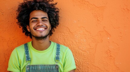 Smiling man with curly hair wearing green t-shirt and denim overalls against orange textured wall.