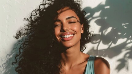 Smiling woman with long curly hair closed eyes and a relaxed expression set against a blurred background with soft shadows and light filtering through.
