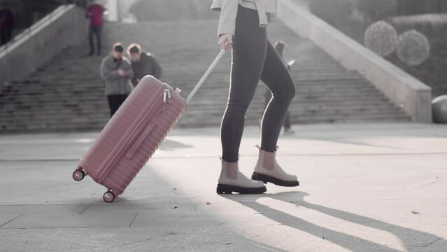 Female tourist walking on a sidewalk in a sunny day and pulling luggage behind her close up, side view. Concept of travel, tourism and enjoying a trip