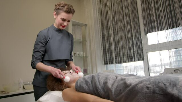 Professional attractive female masseuse doing facial ice globes massage to charming African American woman , reducing puffiness and inflammation , boosting skin elasticity at spa salon