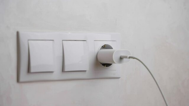 A hand inserts a cell phone charging plug into a wall electrical outlet. Electrical wires and power strips